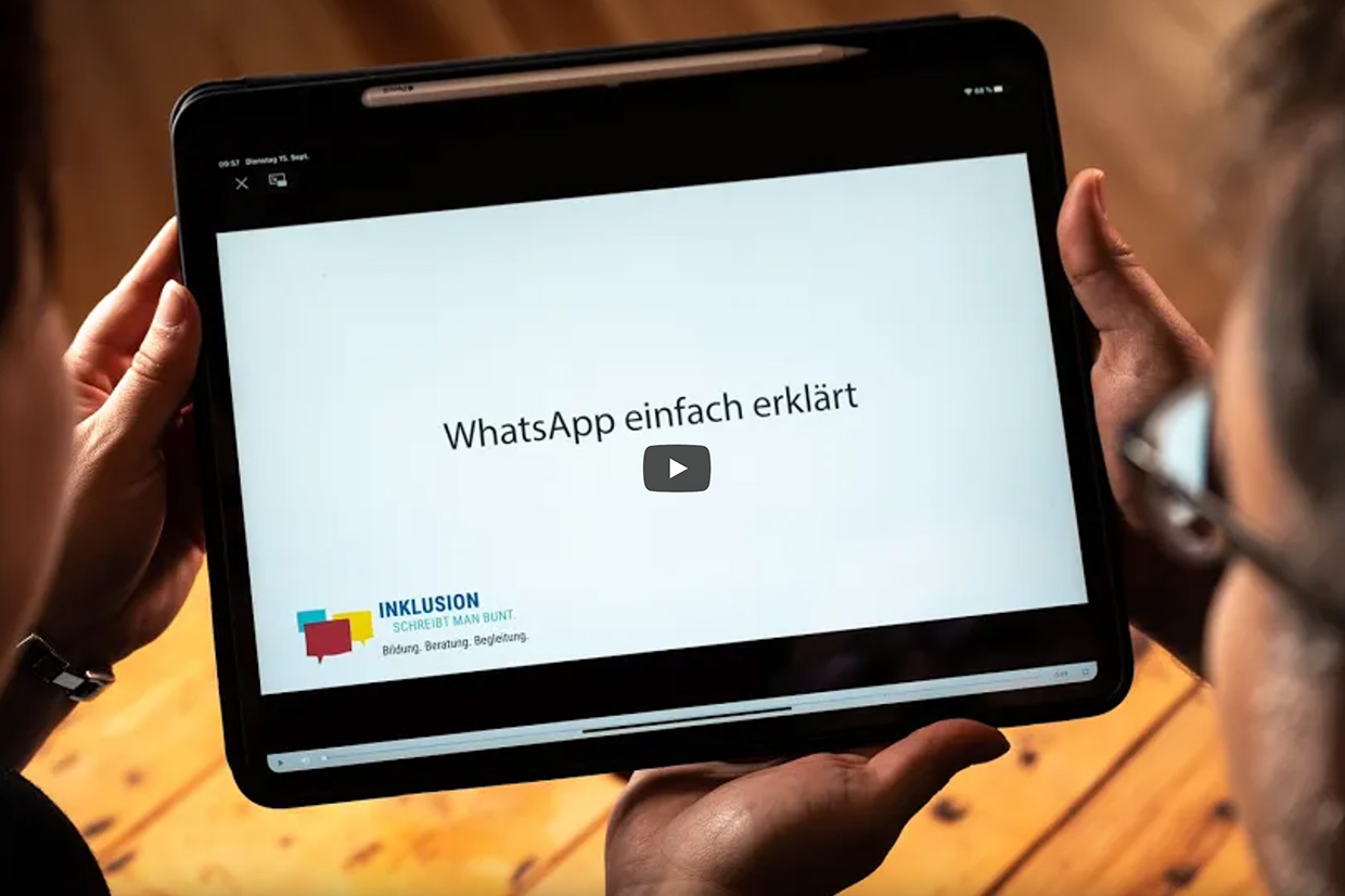 Inklusion_schreibt_man_bunt-WhatsApp-einfach-erklaert Zwei Personen betrachten ein Tablet. Auf dem Tablet ist das Startbild eines YouTube-Videos mit Play-Zeichen in der Mitte zu sehen. Das Video zeigt den Titel